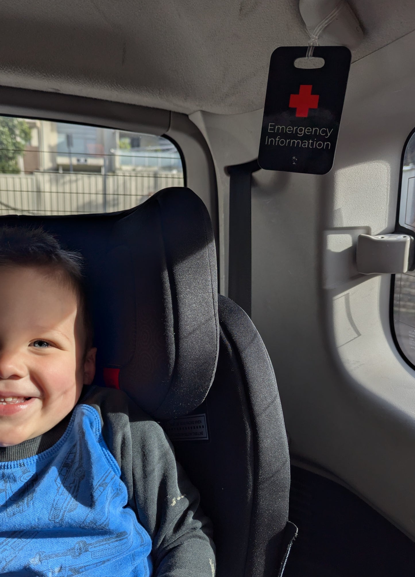 Colour image of Black Hands Here Emergency Medical Information Tag attached to grab handle of car beside forward facing seat with smiling toddler