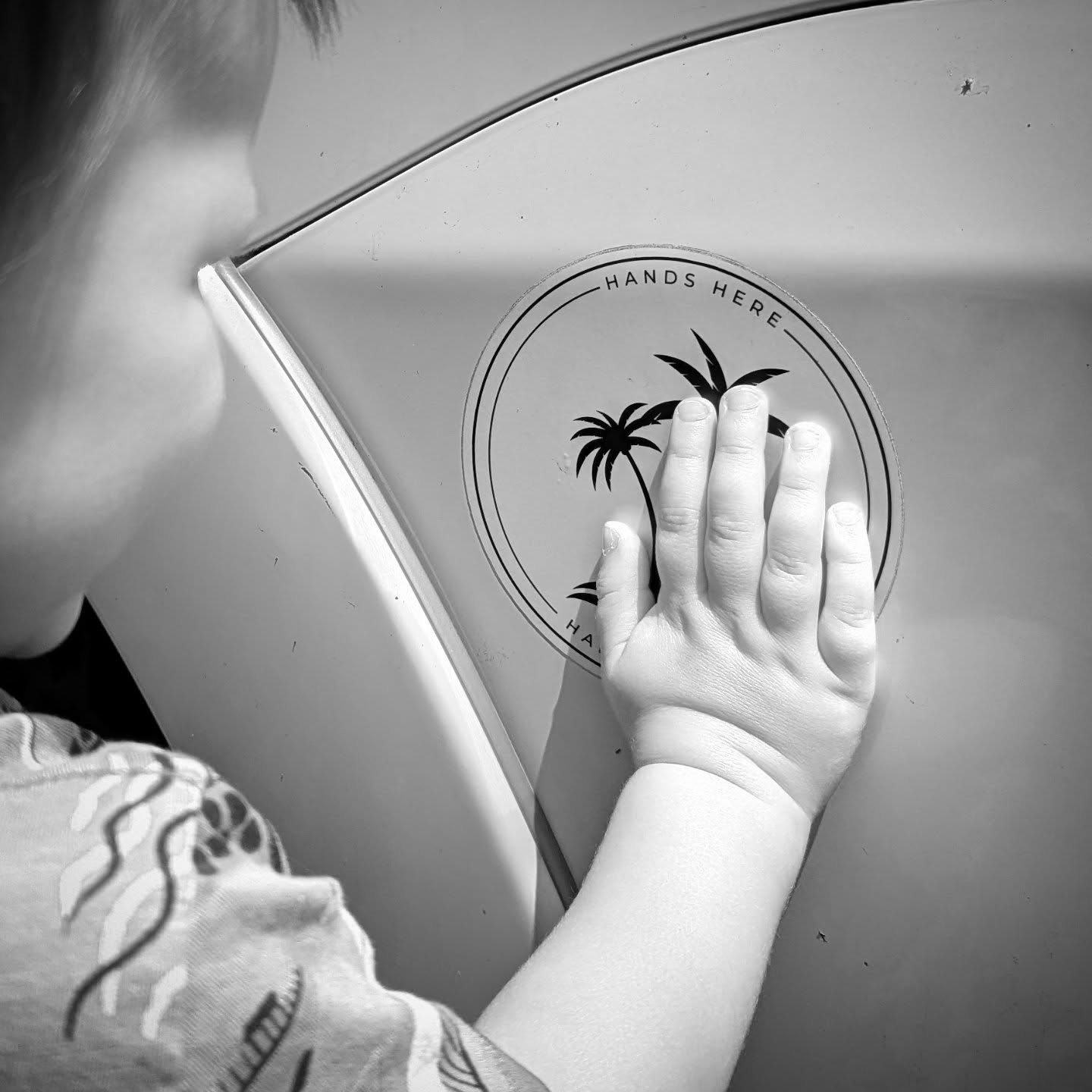 Black and white image of smiling toddler facing car with their hand on the white palm Hands Here decal, safe and stationary near the vehicle.