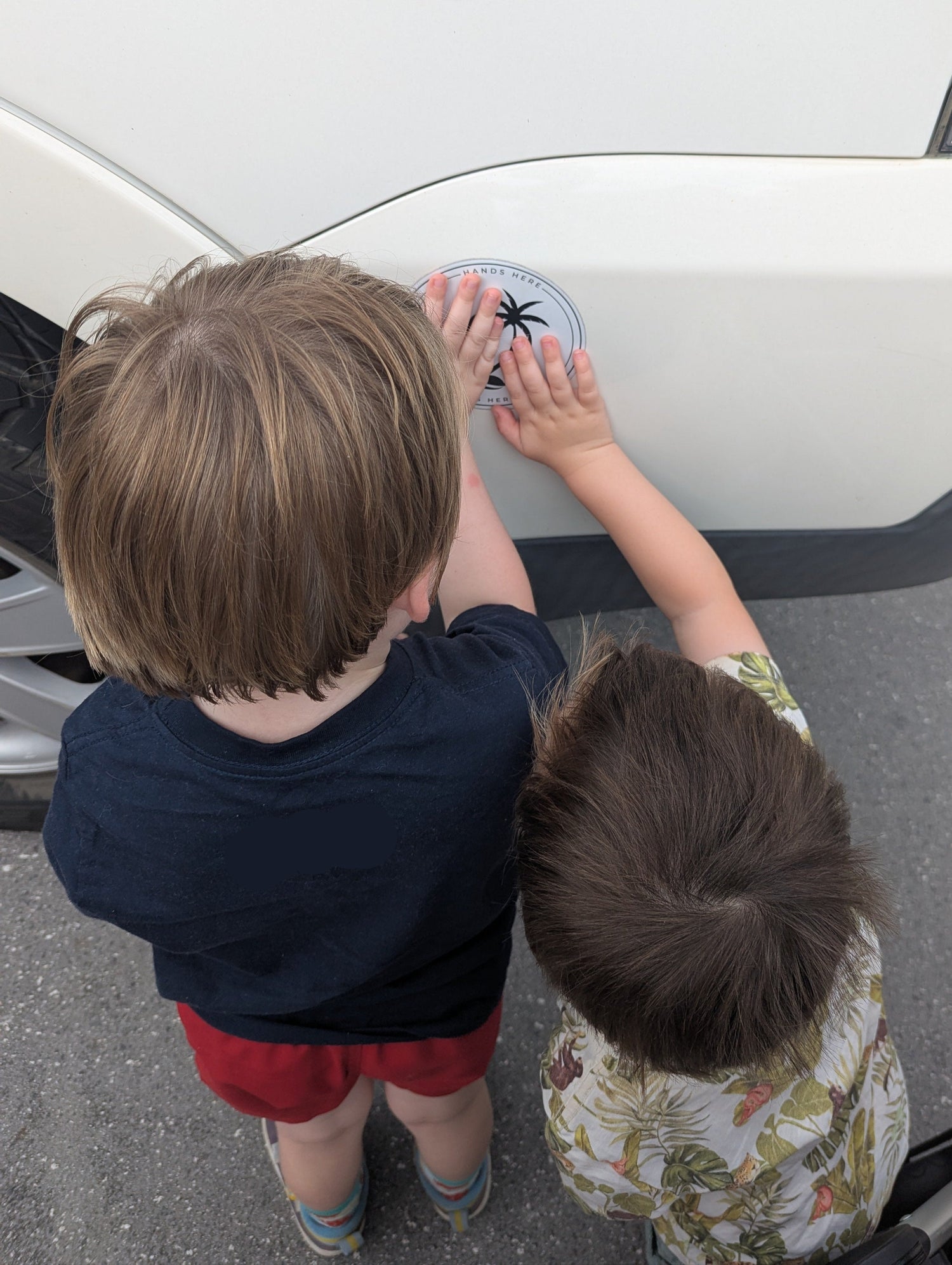 Two toddlers with their hands on the white palm decal on a car facing the vehicle showing them standing stationary and safe.